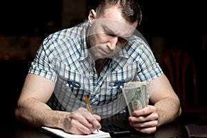 A man with a serious face holds money and takes notes in a notebook