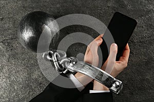 Man shackled with ball and chain holding smartphone at grey table, top view. Internet addiction