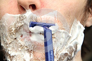 A man shaves his beard with a blue disposable safety razor. White shaving foam on the cheeks and chin