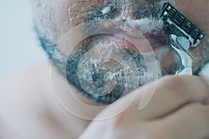 Man is shaving beard his face in bathroom in morning