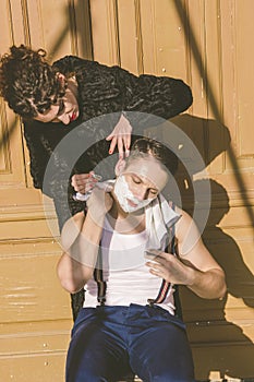 man with shaving foam on his face and towel around his neck siting on chair and posing while pretty women shave him