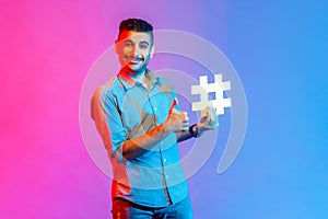 Man in shirt holding hashtag symbol, showing thumb up, recommending popular topics, internet trends.