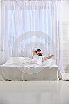 Man in shirt laying on bed, watching tv, white curtains on background. Guy on serious face using remote control for