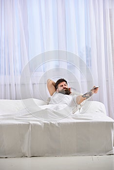 Man in shirt laying on bed, watching tv, white curtains on background. Macho with beard holds controller in hand. Remote