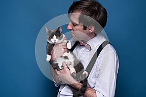 man in shirt, suspender and pink bow tie holding and looking to cute grey cat with white paws on blue background