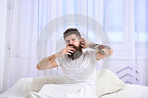 Man in shirt yawning while sit on bed, white curtain on background. Guy on sleepy tired face yawning. Sleepyhead concept