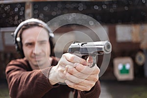 A man shoots a gun, aiming at the target. A man wearing protective headphones. A wall and a roof with bullet holes. outdoor