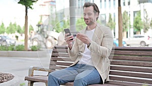 Man Shopping Online via Smartphone while Sitting Outdoor on Bench