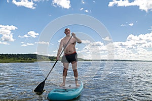 A man in shorts and a T-shirt on a SUP board swims in the lake on a sunny day against a sky with clouds