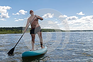 A man in shorts and a T-shirt on a SUP board swims in the lake on a sunny day against a sky with clouds