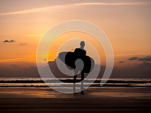 Man silhouette holding surfing board, walking on dark seashore. Surfer standing on sunset sky over the sea water background.
