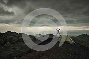 Man silhouette jumping under dramatic sky on volcanic landscape by wild camp in Etna Park