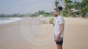 Man sips fresh coconut, strolls along tropical beach. Relaxed male enjoys palm juice on sunny coast. Casual walk