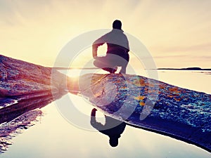 Man sit at evening sea. Hiker sit in squatting position on granite cliff above smooth water.