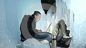 Man is sit on the internet in laptop in an ice cave. Around the mysterious beautiful ice grotto. User communicates in