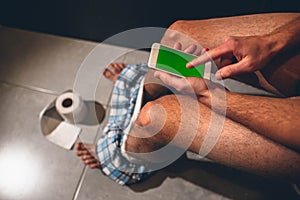Man sit on pot in toilet or rest room. Guy hold phone with green screen and play with it. Point with finger on it and
