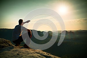 Man sit on rock edge. Hiker makes shadow with hand and watch to colorful mist in forest valley.