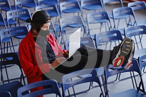 A man sits alone in a large room with many empty blue chairs and works on his laptop.