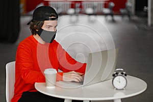 The man sits alone at a table in the open space, drinking coffee and working on his laptop. Freelance concept