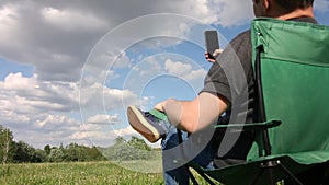 A man sits in a camping chair and photographs the landscape in front of him on the phone. Meadow with green grass. Blue sky with