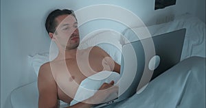 Man sits comfortably soft bed and works intently at laptop. Hands glide easily over keys, bright laptop screen