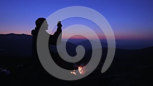 A man sits in the evening by a fire in the mountains and takes pictures of the night landscape on his smartphone. Tourist from