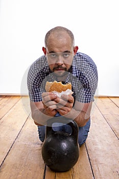 A man sits with a hamburger and a weight on the wooden floor.