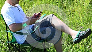 A man sits on a picnic chair in a lighted meadow. He is resting, working on a smartphone. Herbs and flowers sway from the summer