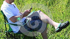 A man sits on a picnic chair in a lighted meadow. He is resting, working on a smartphone. Herbs and flowers sway from the summer w