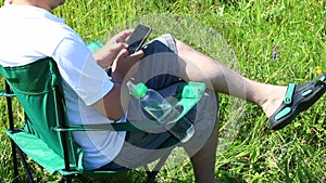 A man sits on a picnic chair in a lighted meadow. He is resting, working on a smartphone. Herbs and flowers sway from the summer w