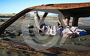 Man sits in rusted car wreck buried in sand on beach