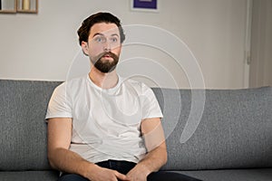 A man sits on a sofa at home, alone, and watches news. He is scared about all. Amazed man reacts to pandemic situation