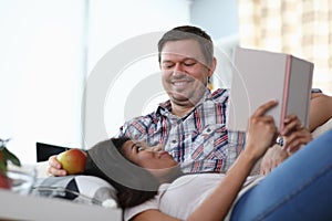 Man sits on sofa smiling on his lap woman lies and holds book in his hands.