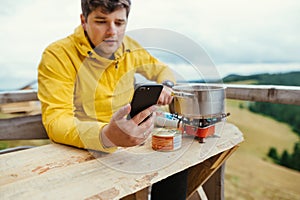 A man sits at a table in the mountains on the terrace of a country house, prepares food on a burner and uses a smartphone