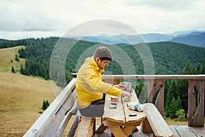 Man sits at a table on a terrace in the mountains and prepares food on a burner on a background of mountain landscape