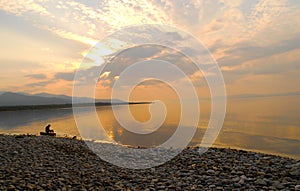 A man sitting alone on a log at the over the edge of a sea, dawn silence on lake.