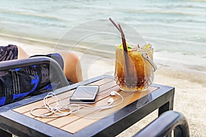 Man sitting at the beach with cocktail bucket, cell phone and earpods on table. Blurred sea in background. Vacation, travel