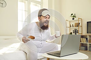 Man sitting on bed, enjoying morning coffee, watching funny movie on laptop and laughing