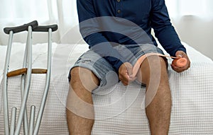 A man sitting on the bed, putting an elastic adhesive bandage on his knee. He leans on crutches, signalling recovery from injury.