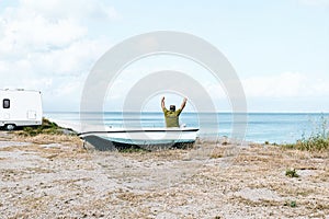 Man sitting in boat on the beach near his white camper van parked by the sea. Tourist season on the mediterranean sea.