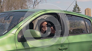 A man sitting in a car and taking a photo with a professional camera, a private detective or a paparazzi spy.