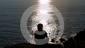 Man sitting on a cliff in front of the sun`s reflection