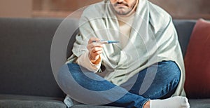 Man Sitting on Couch Covered in Blanket Holding a Thermometer