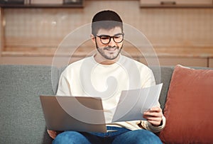 Man Sitting on Couch Examining Paper, Using Laptop