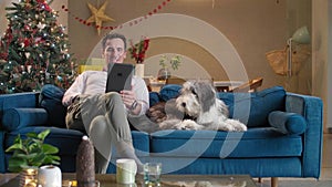 Man is sitting on the couch in living room with Christmas decorations, petting his dog and working on tablet