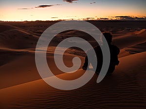 Man sitting on a dune in the desert while watching the sunset.