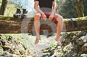Man sitting on the fallen tree log over the mountain forest stream while he waiting for socks laundry drying and trekking boots.