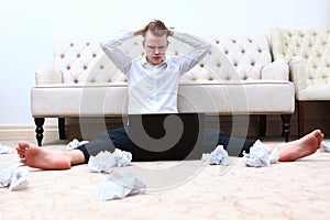 A man is sitting on the floor in front of a laptop. The man is upset and frowns. The financial crisis. Crumpled paper on