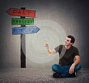 Man sitting on the floor pointing forefinger at signpost arrows shows past, present and future