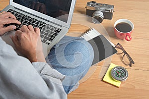 Man sitting on the floor typing on laptop lying on his lap with a cup of coffee,notebook,glasses and camera besides top view.